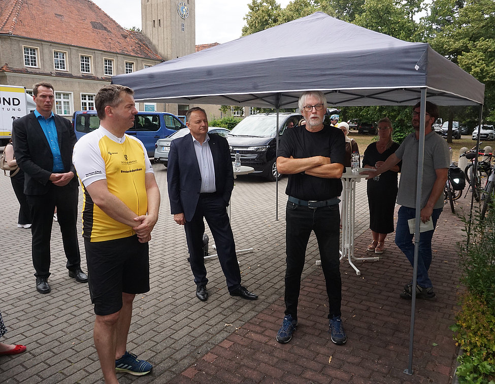 Gruppe von Personen unter einem Pop‑up‑Zelt auf gepflastertem Platz; zwei Männer vorne (einer in gelbem Trikot), weitere Zuhörer und Autos im Hintergrund.